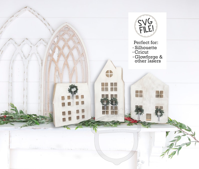 May include: SVG file design of three white wooden houses with wreaths and windows. The houses are on a white shelf with a white background. The text "SVG FILE!" is in a circle at the top of the image. The text "Perfect for: Silhouette, Cricut, Glowforge & other lasers" is below the circle.