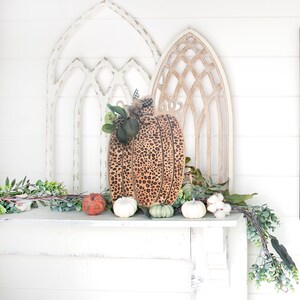 May include: A wooden pumpkin with a leopard print design sits on a white mantel with a garland of greenery and small pumpkins. Two white, one orange, and two green pumpkins are on the mantel.