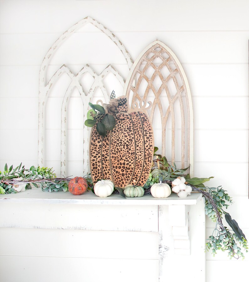 May include: A wooden pumpkin with a leopard print design sits on a white mantel with a garland of greenery and small pumpkins. Two white, one orange, and two green pumpkins are on the mantel.