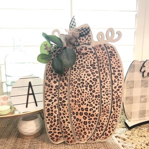 May include: A wooden pumpkin with a leopard print design. The pumpkin is decorated with a burlap bow and green leaves.