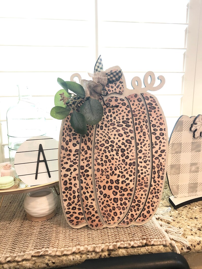 May include: A wooden pumpkin with a leopard print design. The pumpkin is decorated with a burlap bow and green leaves.