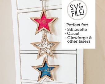 Può includere: Tre stelle di legno con diversi inserti di carta a motivi diversi appesi a una corda. Le stelle sono rosa, bianca e blu. Il testo "SVG FILE!" è in un cerchio nella parte superiore dell'immagine. Il testo sotto il cerchio dice "Perfetto per: Silhouette, Cricut, Glowforge & altri laser".