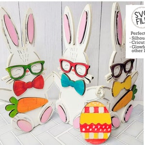 May include: Three wooden bunny figures with glasses and bow ties. The bunnies are white with pink ears and are holding carrots. A red, yellow, and white striped Easter egg is in the foreground.