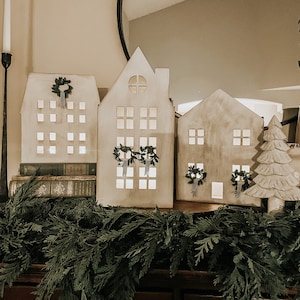 May include: Three white wooden houses with cut-out windows and wreaths on the front, sitting on a wooden surface with a green garland in front of them.