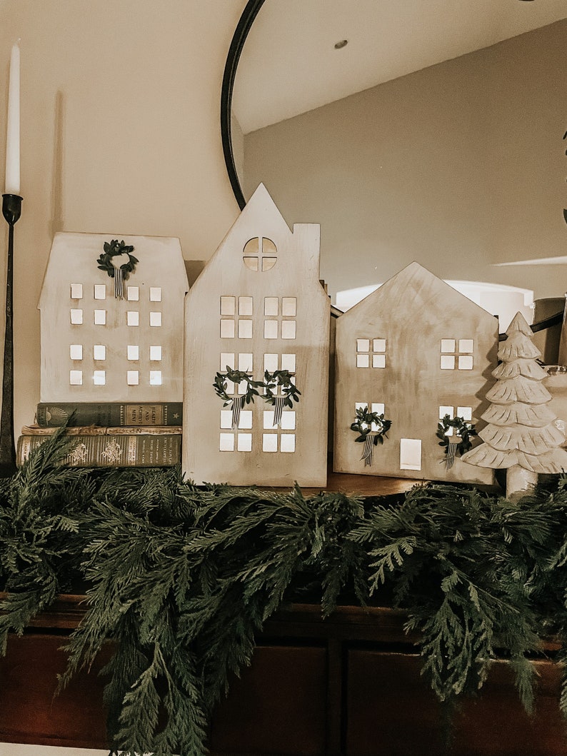 May include: Three white wooden houses with cut-out windows and wreaths on the front, sitting on a wooden surface with a green garland in front of them.