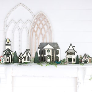 May include: A collection of miniature wooden houses decorated with black and white paint, arranged on a white mantel with greenery. The houses are a church, a house with a wreath, a house with a window, a house with a door, and a house with a sign that says "Fire Dept.".