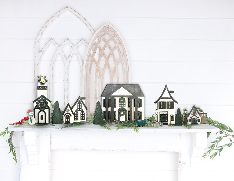 May include: A collection of miniature wooden houses decorated with black and white paint, arranged on a white mantel with greenery. The houses are a church, a house with a wreath, a house with a window, a house with a door, and a house with a sign that says "Fire Dept.".