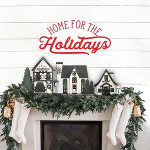May include: A white brick wall with a garland of evergreen branches and red stockings hanging from a fireplace. The garland is decorated with small wooden houses and the text "Home for the Holidays" is written in red letters above the fireplace.