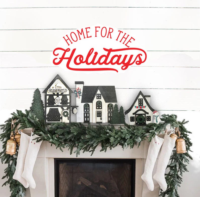 May include: A white brick wall with a garland of evergreen branches and red stockings hanging from a fireplace. The garland is decorated with small wooden houses and the text "Home for the Holidays" is written in red letters above the fireplace.