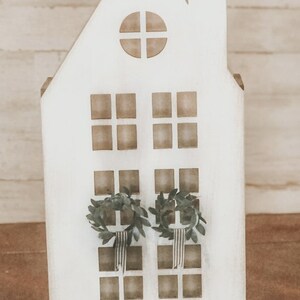 May include: White wooden house shaped ornament with a window and two wreaths hanging on the front. The house is sitting on a brown surface.