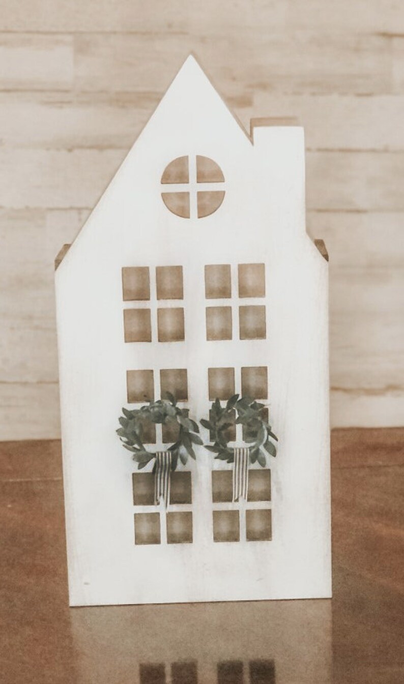 May include: White wooden house shaped ornament with a window and two wreaths hanging on the front. The house is sitting on a brown surface.