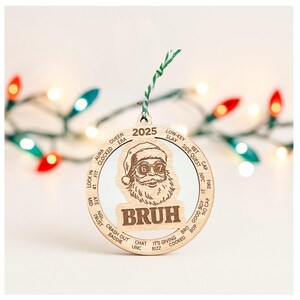 May include: Wooden Christmas ornament featuring a Santa Claus design with sunglasses and the word "BRUH". The ornament has a green and white string for hanging and includes the year 2025. The ornament has various phrases around the edge.