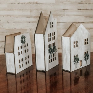 May include: Three white wooden houses with black trim and cut-out windows. Each house has a small wreath with greenery hanging above the front door.