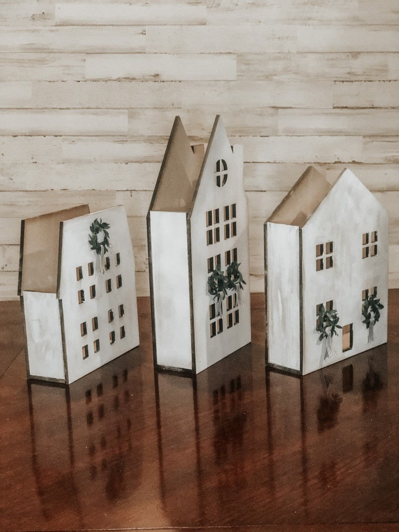 May include: Three white wooden houses with black trim and cut-out windows. Each house has a small wreath with greenery hanging above the front door.