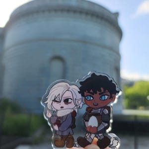 May include: A clear acrylic charm featuring two anime-style characters. One character has white hair and is wearing a scarf and a gray outfit. The other character has dark skin and is wearing armor. The background is a blurred view of a building.