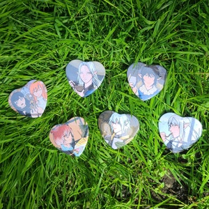 May include: Six heart-shaped pins with different anime characters. The pins are on a green grassy surface.