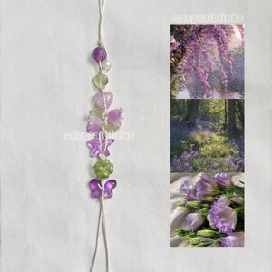 May include: A phone charm featuring a vertical arrangement of purple and green beads in shapes of hearts, flowers, butterflies, and a four-leaf clover on a white cord. To the right, three stacked images show purple wisteria, a bluebell forest, and purple lisianthus flowers with the text adeptalblinks.