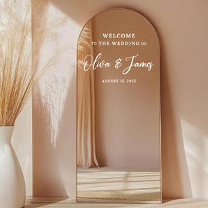 Pode incluir: Um espelho grande e arqueado com moldura dourada. O espelho tem texto branco que diz "WELCOME TO THE WEDDING OF Oliva & James AUGUST 15, 2025". O espelho está numa sala com uma parede bege e um vaso branco com plantas secas.