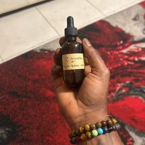 May include: A brown glass bottle of citronella oil with a black dropper top. The label reads "Citronella oil, dilute before use." The bottle is held in a hand, with two beaded bracelets on the wrist.