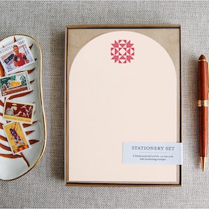 May include: A stationery set with a light pink design, a brown pen, and a decorative dish with stamps. The stationery set includes a stack of light pink paper with a red geometric design. The pen is brown with gold accents.