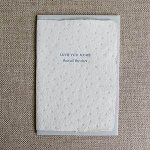May include: A white card with a blue embossed message that reads "Love you more than all the stars". The card is decorated with a subtle pattern of silver stars.