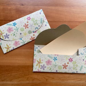Könnte beinhalten: Zwei Briefumschläge aus Papier mit floralem Muster. Einer ist geschlossen, der andere ist offen mit einer hellbraunen Karte darin. Die Umschläge haben einen cremefarbenen Hintergrund mit bunten Blumen und Libellen.
