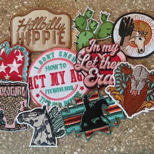 May include: A collection of embroidered patches with various designs and text. The patches feature a cowgirl, a horse, cacti, and phrases like "Hillbilly Hippie" and "In My Let the Era". The patches are in various shapes and colors.