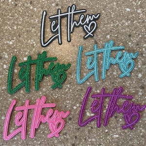 May include: Five glittery word art pieces spelling "Let them" with a heart. The colors are black and white, green, teal, pink, and purple. The words are in a cursive font.