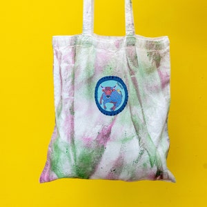 May include: White tote bag with green and pink tie dye pattern. The bag has a blue circle patch with a red bull inside and the word "Taurus" written on the patch.