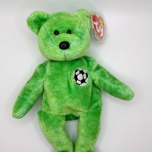 Puede incluir: Un oso de peluche verde con un parche de balón de fútbol blanco y negro en el pecho. El oso tiene una nariz negra y ojos negros pequeños. Una etiqueta roja y blanca está sujeta a su oreja.
