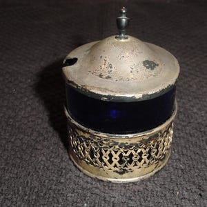 May include: A vintage condiment jar featuring a deep cobalt blue glass body encased in a tarnished silver-tone metal filigree holder. The jar includes a matching silver-tone metal lid with a small finial handle and a side notch for a spoon.