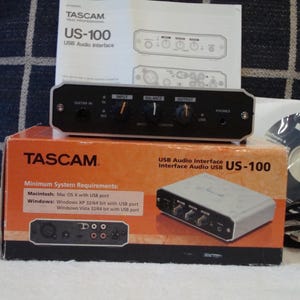 Puede incluir: Interfaz de audio USB TASCAM US-100. La interfaz negra y plateada se encuentra sobre una caja naranja y negra. La caja incluye un CD-ROM, cables y un manual de usuario. La interfaz tiene controles de entrada, balance y salida.