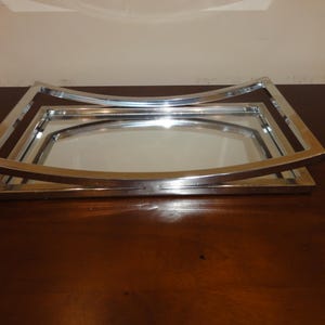 Puede incluir: Una bandeja rectangular de metal plateado con una base de espejo y un marco elevado y curvado. La estructura metálica pulida crea una apariencia de capas con una superficie reflectante que refleja el entorno circundante.
