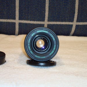 May include: A Quantaray camera lens with a black body and a lens cap. The lens has the following specifications: 1:4-5.6, f=70-210mm, and a 55mm diameter. The lens is multi-coated and made in Japan.