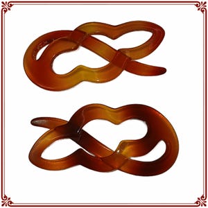 May include: Two amber-colored hair clips in a figure-eight design. The clips have a glossy finish and a gradient of colors, transitioning from a lighter yellow to a darker brown. The design is a continuous loop, creating a stylish accessory.