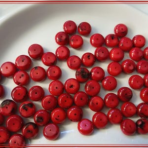 May include: A collection of round, bright red beads with a glossy finish. Each bead has a small hole in the center. Some beads have dark spots. The beads are scattered on a white surface, likely for jewelry making.