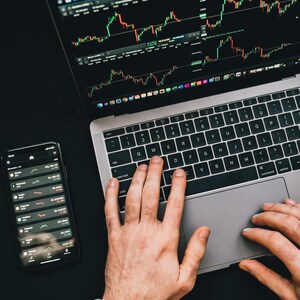 May include: A person's hands are typing on a laptop computer with a stock market chart displayed on the screen. A smartphone with a stock market app is also visible on the table.