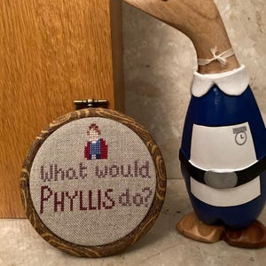 Pode incluir: Uma moldura redonda de madeira com um fundo de tecido bege e bordado vermelho que diz "What would Phyllis do?" com uma pequena imagem bordada de uma mulher com uma jaqueta vermelha.