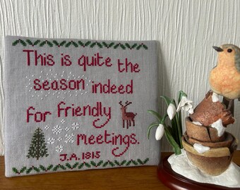 Op de afbeelding: Een kruissteek met de tekst "This is quite the season indeed for friendly meetings." Een kleine kerstboom en een hert zijn ook op de grijze stof geborduurd. Er staat een keramieken vogelbeeldje naast.
