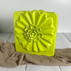 May include: A square, lime green ceramic vase featuring a prominent, three-dimensional sculpted flower design on the front. The flower has layered, ruffled petals that radiate from the center, creating a textured, glossy surface finish.