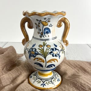 May include: A ceramic vase with a scalloped rim and two curved handles. The white surface features hand-painted floral motifs in shades of blue, yellow, and green. The base is circular with a yellow band and decorative blue and green leaf patterns.
