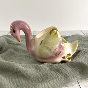 May include: A ceramic swan-shaped planter or dish featuring a glossy finish with blended pastel pink and cream tones. The swan has gold-painted accents on its head, neck, and wings, with a curved neck and an open cavity on its back for storage.