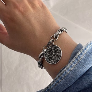 Puede incluir: Un brazalete de cadena de plata con un gran colgante circular que presenta intrincados diseños negros y letras.