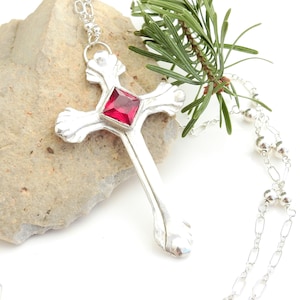 Könnte beinhalten: Eine silberne Halskette mit einem roten Edelstein-Anhänger in Kreuzform. Die Halskette liegt auf einem weißen Hintergrund mit einem Stein und einem Tannenzweig.