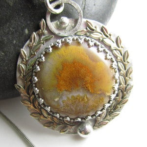 May include: A silver pendant featuring a round, yellow and white agate stone set within a silver frame. The frame is adorned with a silver leaf design.