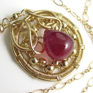 Könnte beinhalten: Goldfarbene Anhängerkette mit einem großen, tropfenförmigen, tiefroten Edelstein. Der Edelstein ist in einem kreisförmigen, drahtgewickelten Design mit kleinen goldenen Perlen gefasst. Die Halskette ist ebenfalls goldfarben.