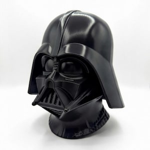 May include: A black Darth Vader helmet, a bust of the Star Wars character. The helmet has a glossy finish and detailed facial features, including the mask, respirator, and helmet shape. The bust is a solid, three-dimensional object.