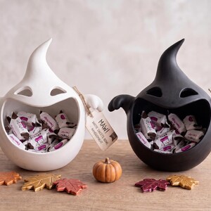 Può includere: Due ciotole a forma di fantasma, una bianca e una nera, riempite con caramelle incartate singolarmente. Ogni ciotola ha un'etichetta con scritto MAW! WHITE SWEETS BOWL - HANDGEMACHT o MAW! BLACK SWEETS BOWL - HANDGEMACHT. Una piccola zucca e cinque foglie autunnali sono poste davanti.