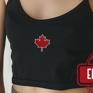 May include: A black cropped tank top featuring a small, centered red maple leaf embroidery. The garment has thin shoulder straps and a scoop neckline. A red circular stamp with the text EMBROIDERED is visible in the lower right corner.