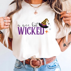 May include: Cream-colored t-shirt with the phrase "a wee bit WICKED" in black and purple text. A witch's hat with black, yellow, and purple accents is featured above the word "WICKED", with star and orange accents.